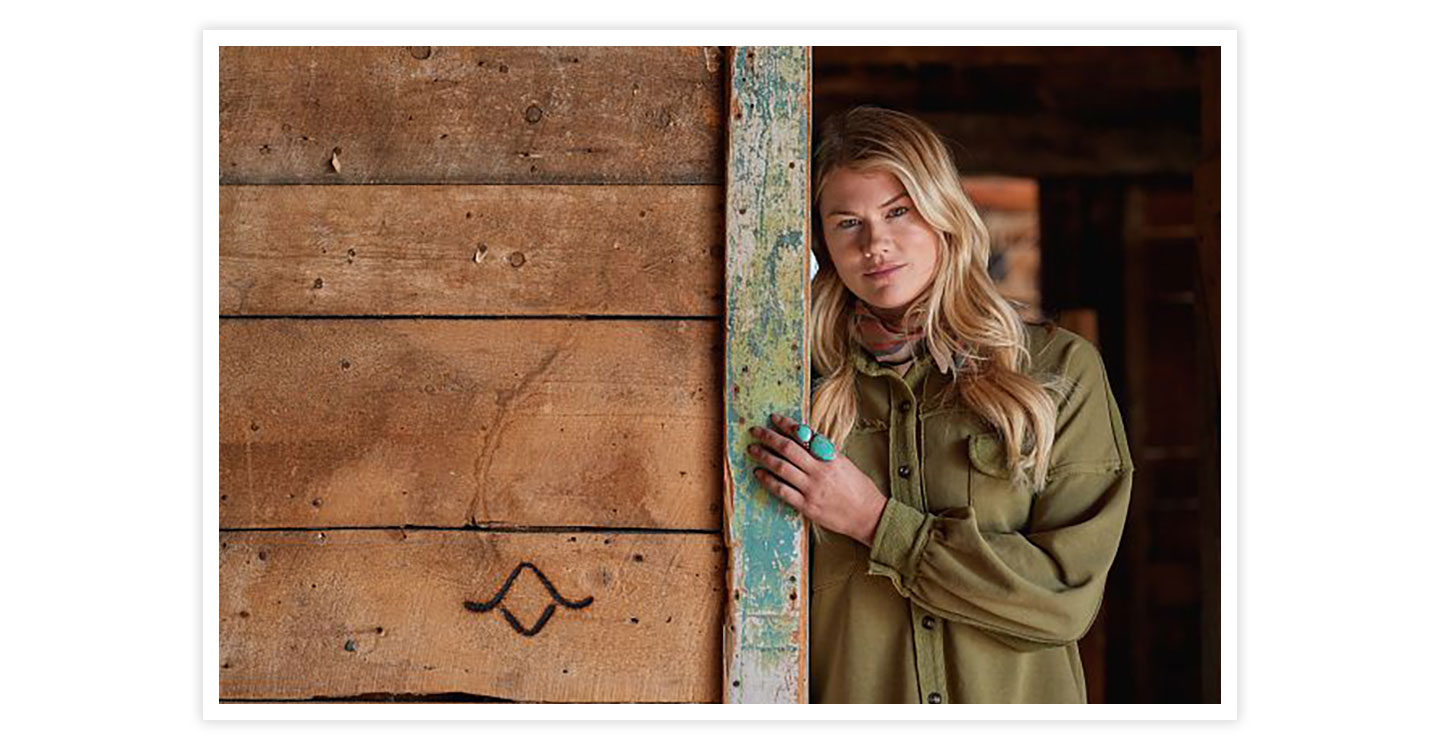 A woman leaning against a brown wood barn door wear a green button-down shirt and turquoise jewelry.